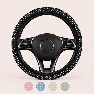 May include: A black steering wheel cover featuring a white polka dot pattern. The cover is fitted onto a black steering wheel with integrated control buttons. Below the steering wheel are four circular color swatches in pink, blue, beige, and green.