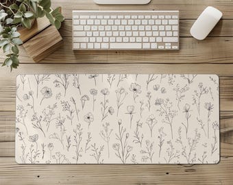 Puede incluir: Alfombrilla de escritorio rectangular con fondo crema y delicado estampado floral en gris oscuro. El diseño presenta varias flores y follaje esbozados, creando un tema botánico. La alfombrilla está colocada sobre un escritorio de madera.