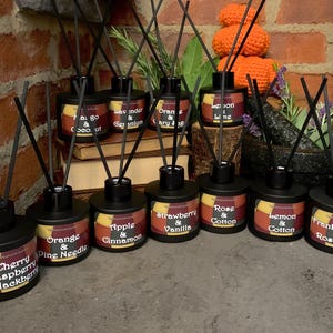 May include: A collection of ten black glass diffuser bottles with various scents, including cherry raspberry & blackberry, orange & pine needle, apple & cinnamon, strawberry & vanilla, rose & cotton, lemon & cotton, frankincense & rosewood, mango & coconut, lavender & geranium, and orange & cherry. The bottles are arranged in a row on a brown surface.