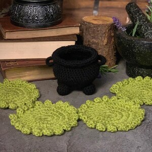 May include: Four green crocheted coasters with a black crocheted cauldron in the centre. The cauldron is sitting on a stack of books and a wooden log.