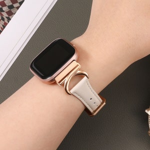 Könnte beinhalten: Ein weißes Lederarmband mit einer roségoldenen Schließe und einem schwarzen Zifferblatt. Das Armband wird am Handgelenk einer Person getragen.