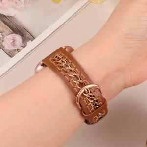 Könnte beinhalten: Braunes Leder-Uhrenarmband mit dekorativem Kettenmuster. Das Armband wird mit einer goldfarbenen Schnalle befestigt. Das Uhrenarmband wird an einem Handgelenk gezeigt.