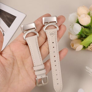 以下が含まれることがあります： 銀色のバックルが付いたベージュの革製の腕時計バンド2本。右側のバンドには、ハートマーク付きの「M&C」のイニシャルが刻印されています。