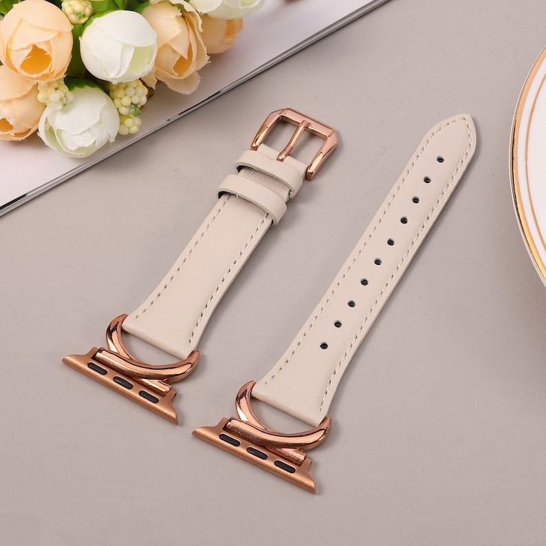 Puede incluir: Una correa de reloj de cuero blanco con herrajes de oro rosa. La correa tiene un cierre de hebilla y un dise&ntilde;o de doble bucle.