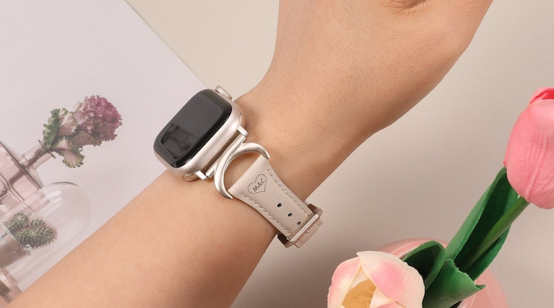 Puede incluir: Una correa de reloj de cuero blanco con una hebilla plateada y un dise&ntilde;o de media luna. La correa tiene las iniciales "M&C" grabadas con un coraz&oacute;n.
