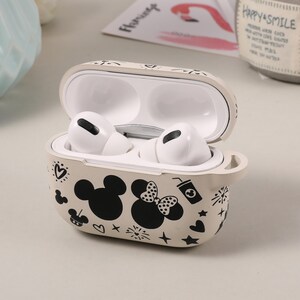 May include: A beige protective case for wireless earbuds with a black and white design featuring Mickey Mouse and Minnie Mouse. The case has a small loop on the side.