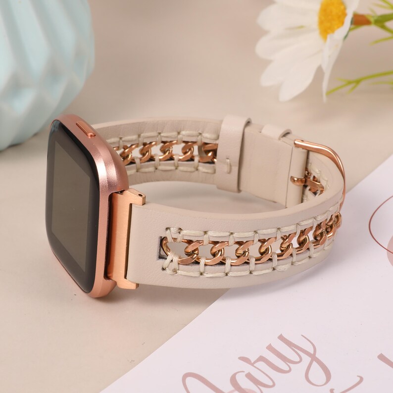 May include: A rose gold smartwatch with a cream-colored leather band. The band features a gold chain design woven into the leather. The watch is displayed on a light-colored surface.