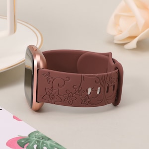 May include: A rose gold smartwatch with a maroon silicone band featuring a floral design. The band has a detailed floral pattern with leaves and flowers.