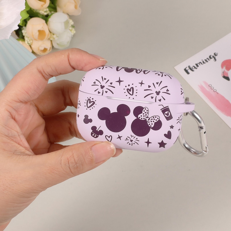 May include: Purple silicone case for wireless earbuds with a Mickey Mouse and Minnie Mouse design. The case has a black and white print of fireworks, stars, hearts, and a cup. The case has a silver carabiner.