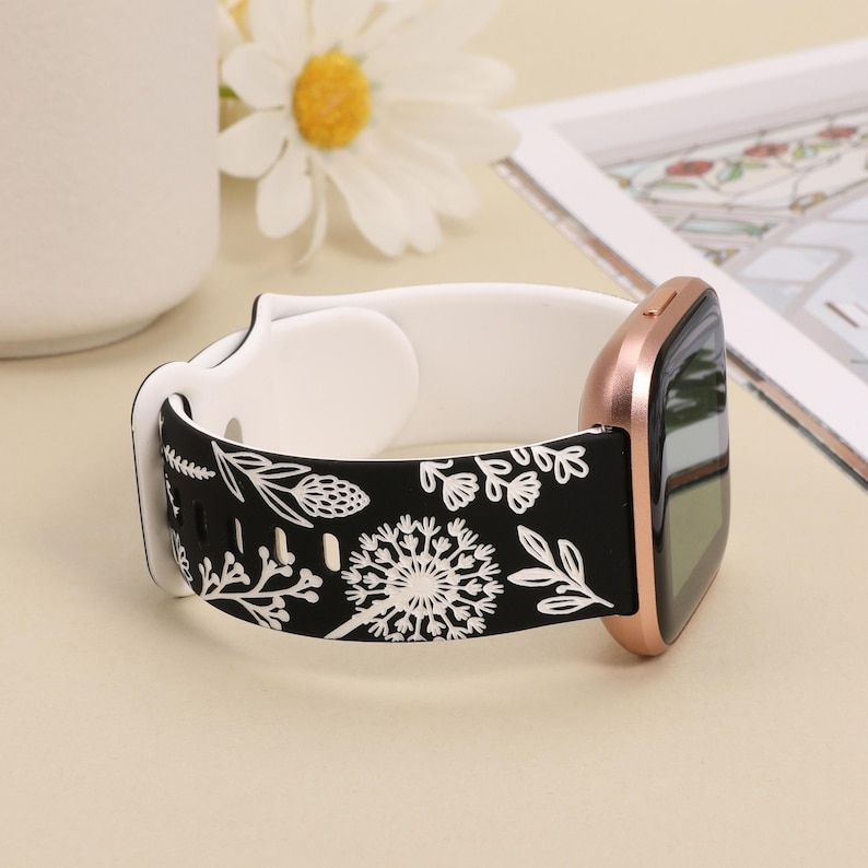 May include: Black and white floral patterned watch band with a rose gold watch face. The band is made of silicone and has a white buckle.