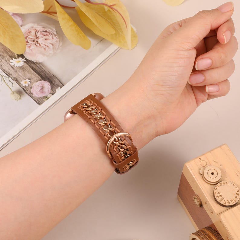 May include: A brown leather watch band with a decorative chain pattern. The band is secured with a gold-colored buckle. The watch band is shown on a wrist.