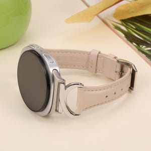 May include: A silver smartwatch with a black screen and a light beige leather band. The band has a silver buckle and is attached to the watch with silver hardware. The watch is on a light beige surface.