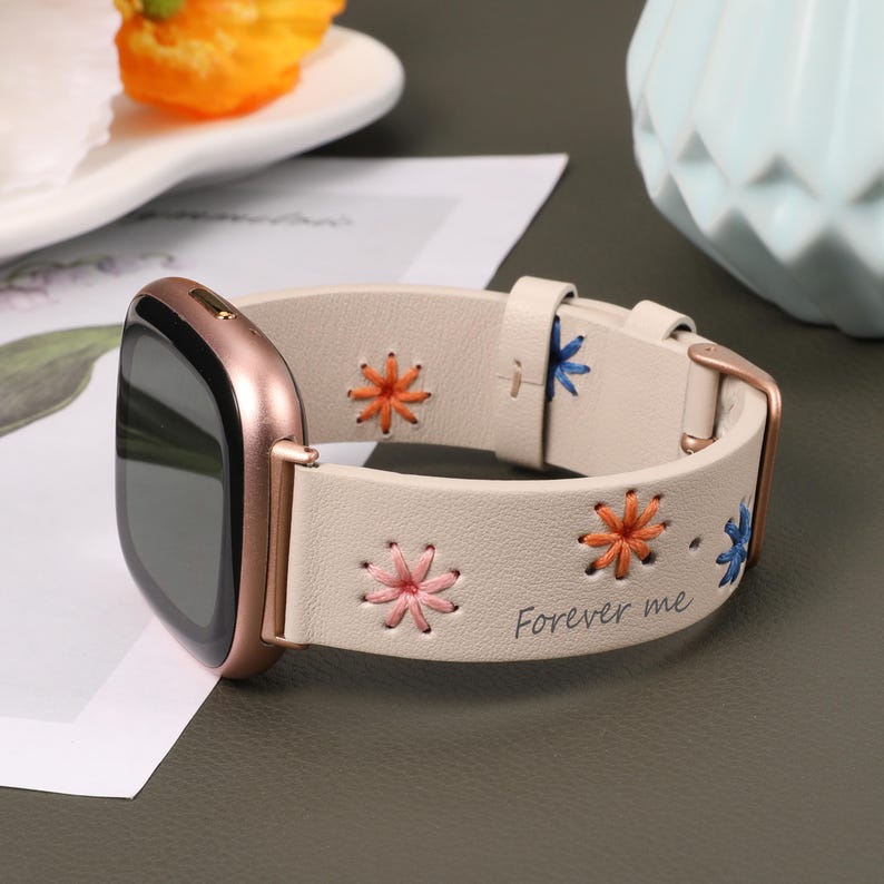Puede incluir: Una correa de reloj de cuero beige con flores bordadas de colores y el texto "Forever me".