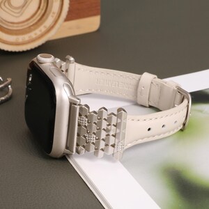 May include: A silver Apple Watch with a white leather band and a silver metal band with a geometric pattern. The band is labeled "genuine leather".