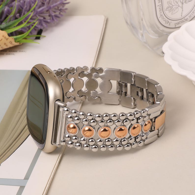 Puede incluir: Un reloj inteligente con caja en tono dorado y una pulsera de eslabones m&uacute;ltiples. La pulsera presenta un patr&oacute;n repetitivo de cuentas plateadas pulidas y detalles circulares en tono oro rosa, con una apariencia texturizada.