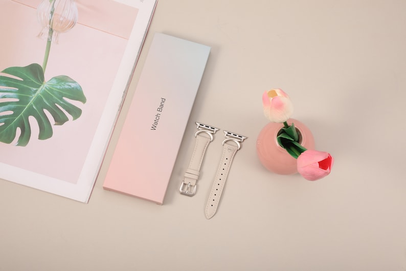 Puede incluir: Dos correas de reloj de cuero beige claro con cierres y hebillas plateadas. Las correas est&aacute;n dispuestas sobre una caja de degradado rosa claro y azul que dice "Watch Band".