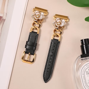 May include: Two black leather watch straps with gold-tone hardware. Each strap features a gold-tone chain and a decorative floral accent with white petals and clear crystals. The straps are designed for a watch.