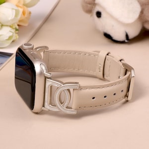 May include: A silver Apple Watch with a cream-colored leather band. The band features a silver buckle and a decorative silver clasp. The watch is displayed on a light-colored surface.