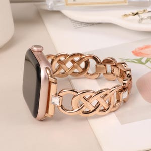 以下が含まれることがあります： ローズゴールドのアップルウォッチバンド。編み込みのデザインで、クリアなクリスタルがアクセントになっている。