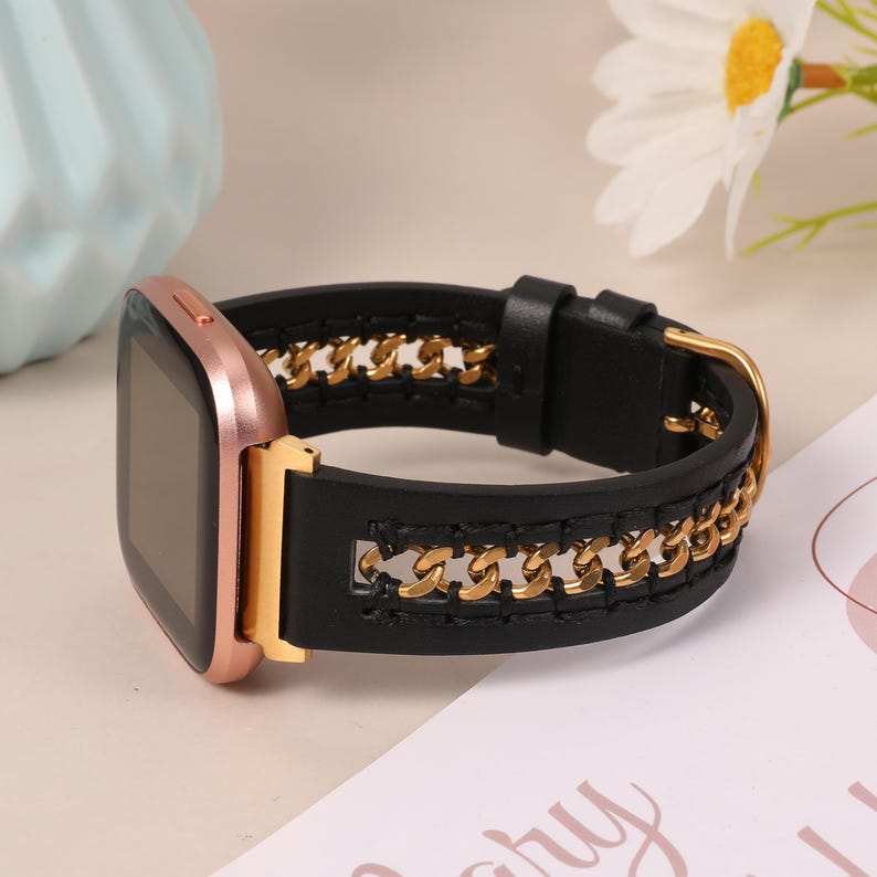 May include: A rose gold smartwatch with a black leather band featuring a gold chain design. The watch has a rectangular screen and a gold buckle. The band is connected to the watch with gold hardware.