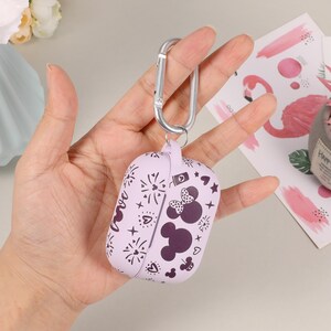 May include: A purple silicone case with a Minnie Mouse design and a silver carabiner clip. The case has a pattern of fireworks, hearts, and stars. The case is designed to fit AirPods.