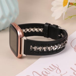 May include: A rose gold smartwatch with a black leather band featuring a silver chain design. The watch has a square face and a buckle closure. The band is approximately 1 inch wide.