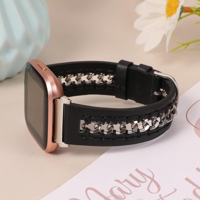 May include: A rose gold smartwatch with a black leather band featuring a silver chain design. The watch has a square face and a buckle closure. The band is approximately 1 inch wide.