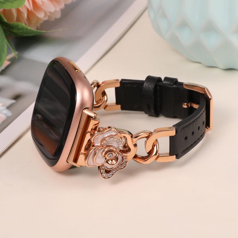 May include: Rose gold smartwatch with a black leather band. The band features a decorative rose with white petals and crystal accents. The watch case is rose gold, and the band is connected with a chain-link design.