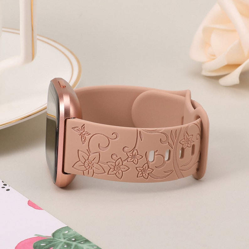May include: A rose gold smartwatch band with a floral design. The band is made of silicone and has a rose gold buckle.
