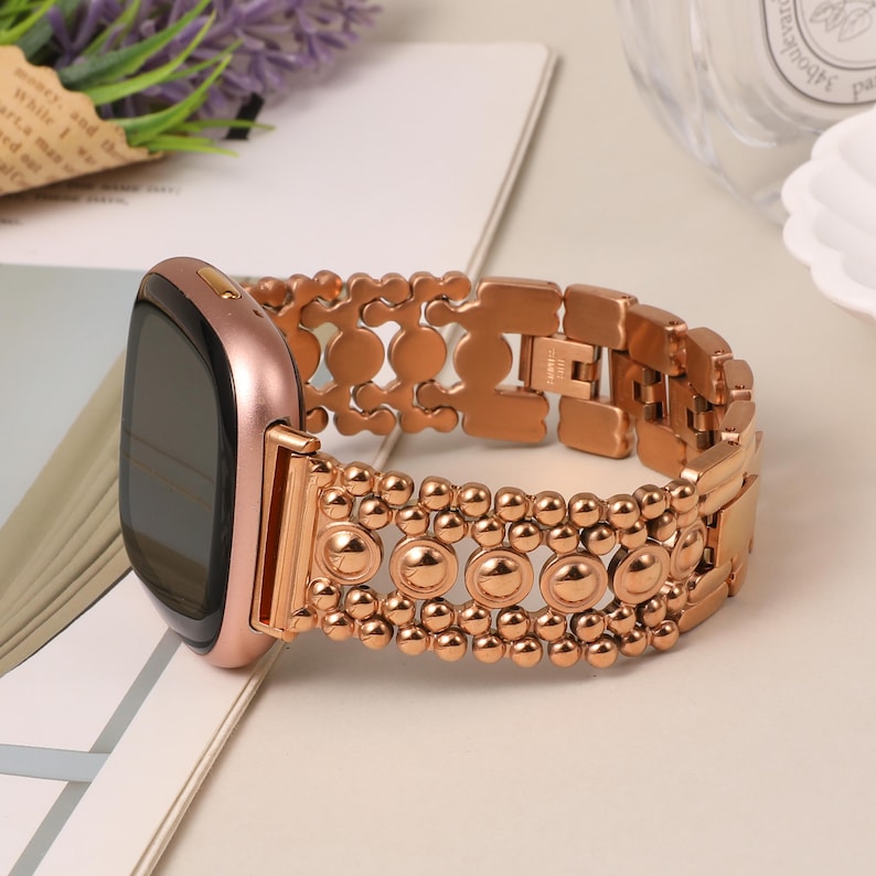 Puede incluir: Un reloj inteligente en oro rosa con una correa de eslabones met&aacute;licos que presenta un patr&oacute;n decorativo de peque&ntilde;as esferas y detalles circulares. La caja del reloj es redondeada, con pantalla negra y acabado pulido en tono oro rosa.