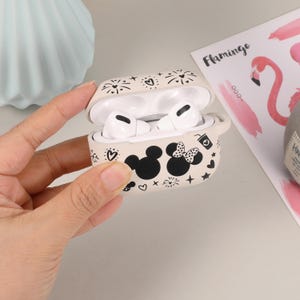 May include: A white silicone case for wireless earbuds with a black and white design featuring Mickey Mouse and Minnie Mouse. The case is open and the earbuds are visible inside.