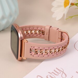 May include: A rose gold smartwatch with a pink leather band. The band features a gold chain design woven into the pink leather. The watch has a square face and a gold buckle.