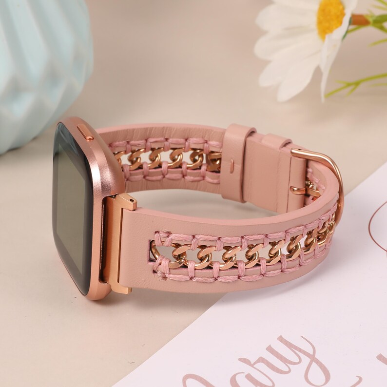 May include: A rose gold smartwatch with a pink leather band. The band features a gold chain design woven into the pink leather. The watch has a square face and a gold buckle.