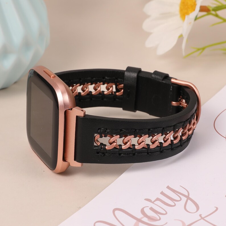 May include: Black leather watch band with rose gold chain accents. The watch band is attached to a rose gold smartwatch. The band features a chain design and a buckle closure. The watch face is black.