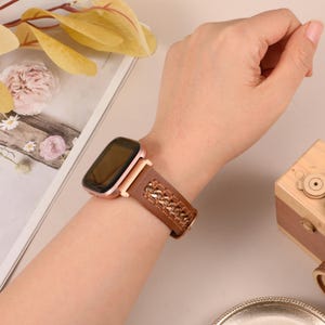 May include: A rose gold smartwatch with a brown leather band featuring a woven design is worn on a wrist. The watch face is rectangular with a black screen. The band is secured with a rose gold clasp.