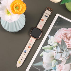 Puede incluir: Una correa de reloj de cuero blanco con flores bordadas de colores y las iniciales "JM".