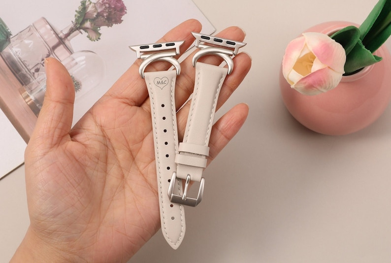 Puede incluir: Dos correas de Apple Watch de cuero beige con hebillas plateadas y un coraz&oacute;n plateado con las iniciales "M&C" grabadas en una correa.