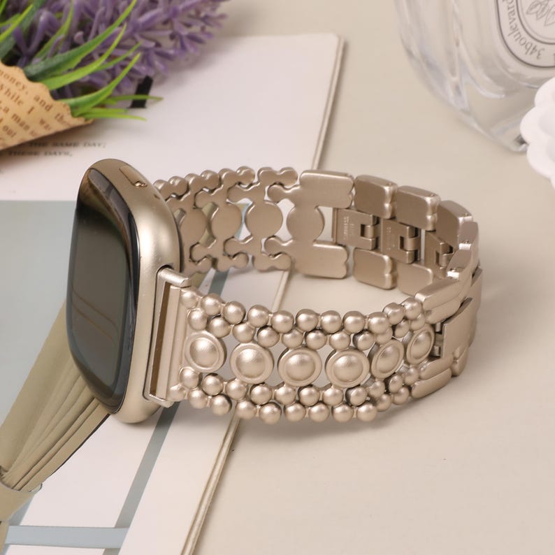 Puede incluir: Un reloj inteligente color champ&aacute;n con una correa de eslabones met&aacute;licos que presenta un dise&ntilde;o decorativo de peque&ntilde;as esferas y detalles circulares. La caja del reloj es lisa y redondeada, a juego con la pulsera de metal texturizada.