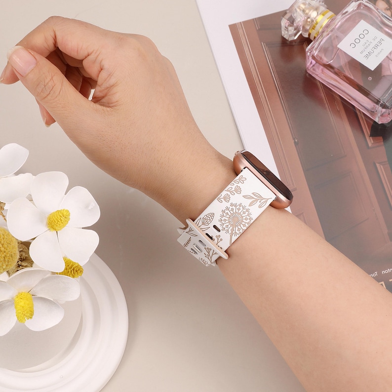May include: White silicone watch band with a floral design in gold. The band is compatible with Apple Watch.