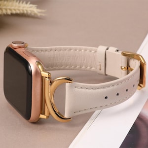Puede incluir: Un Apple Watch de oro rosa con una correa de cuero blanco y una hebilla dorada. La correa tiene un cierre de anillo en D.