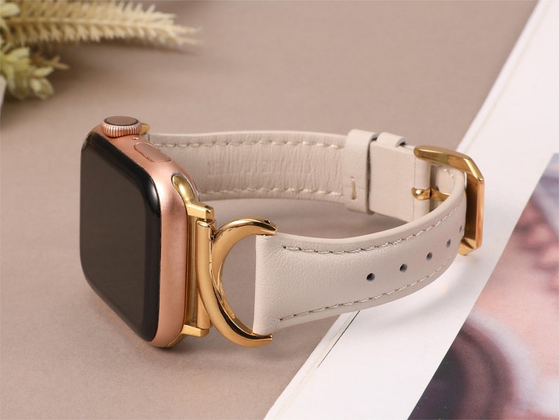 Puede incluir: Un Apple Watch de oro rosa con una correa de cuero blanco y una hebilla dorada. La correa tiene un cierre de anillo en D.