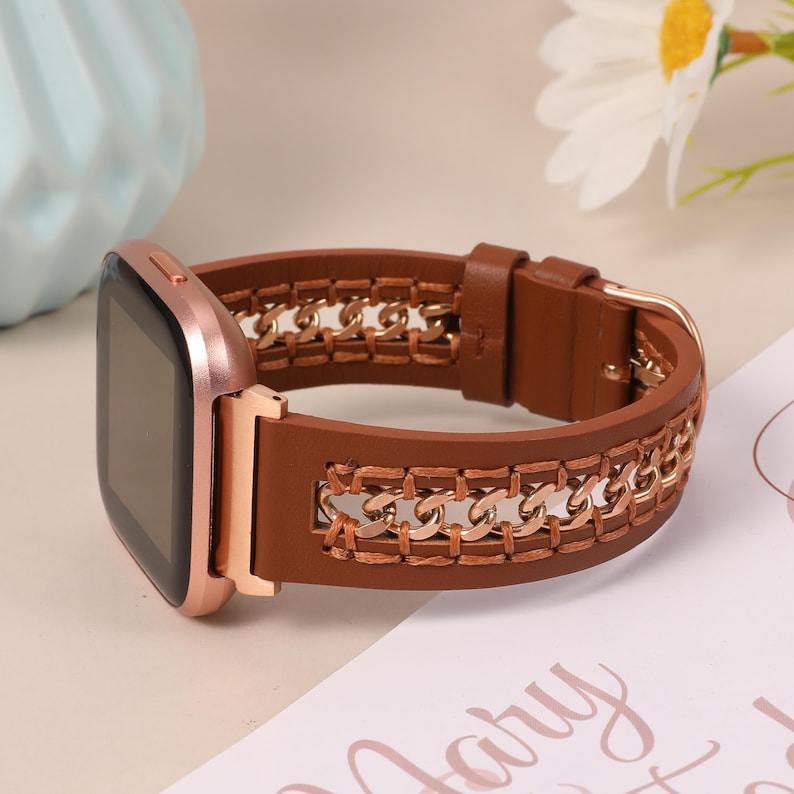 May include: Brown leather smartwatch band with gold chain detailing. The band features a rose gold watch face and a gold buckle. The watch band is a stylish accessory for a smartwatch.