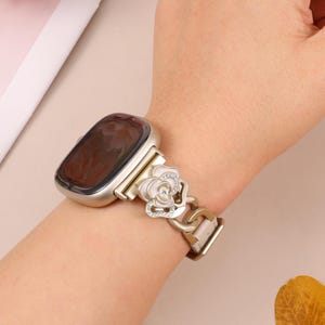 May include: A silver smartwatch with a light pink band. The band features a decorative flower with white petals and sparkling rhinestones. The watch is worn on a wrist.