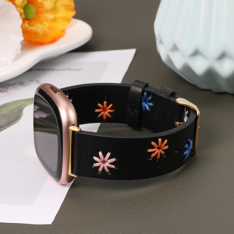 Puede incluir: Una correa de reloj de cuero negro con flores bordadas de colores. La correa es compatible con relojes inteligentes.