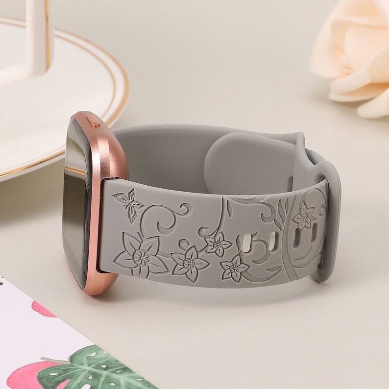 May include: A gray silicone watch band with a floral design. The band is compatible with Fitbit Versa smartwatches.