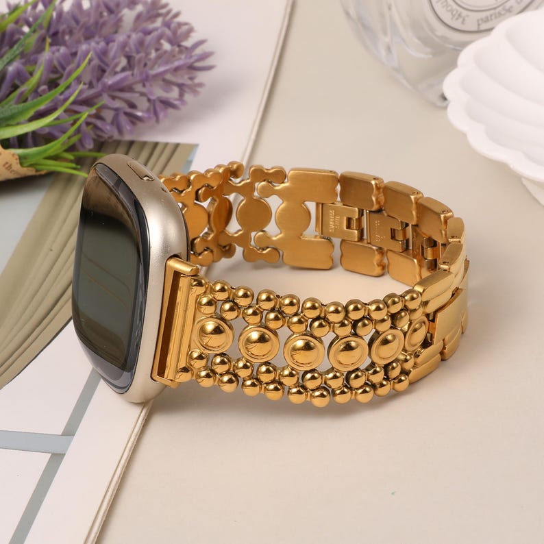 Puede incluir: Una correa de reloj inteligente en tono dorado con un dise&ntilde;o de eslabones decorativos que incluye peque&ntilde;as esferas pulidas y detalles circulares. La correa est&aacute; unida a una caja de reloj met&aacute;lica con pantalla oscura y dise&ntilde;o estructurado.