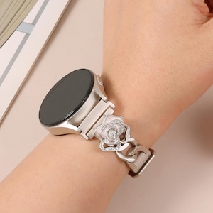 Op de afbeelding: Een zilverkleurige smartwatch met een decoratieve band. De band heeft een bloemmotief met een roosvormig middelpunt versierd met kleine, sprankelende details. De wijzerplaat van het horloge is zwart en rond, en de band is vastgezet met een sluiting.