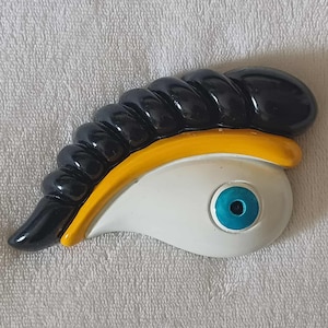 Puede incluir: Objeto decorativo en forma de ojo estilizado. El diseño presenta una parte superior negra, brillante y curvada, una banda amarilla y un ojo blanco con iris azul. El objeto probablemente está hecho de cerámica o un material similar.