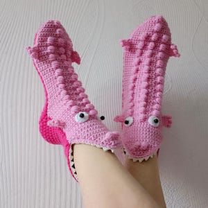 May include: Pink crocheted alligator slippers with white eyes and teeth. The slippers are worn on a person's feet.