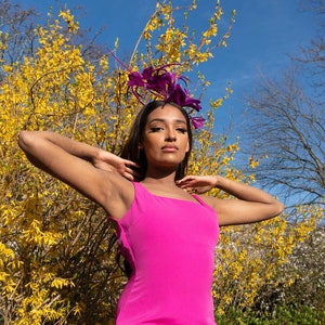 May include: A woman wearing a bright pink sleeveless dress with a purple flower hat. She is standing in front of a bush with yellow flowers.